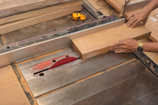 Close-up of hands guiding a wooden board through a table saw with a red blade, used for precise straight cuts in woodworking.