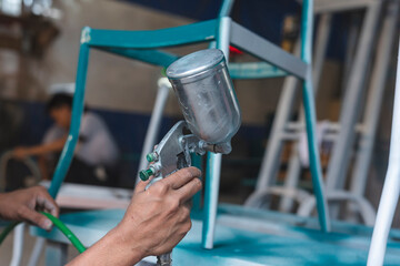 Worker using a spray gun to apply teal paint on a steel or iron chair frame during furniture...