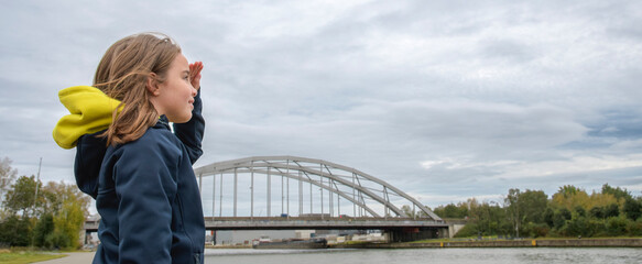 In front of a large iron bridge across the river, a girl stares along the horizon