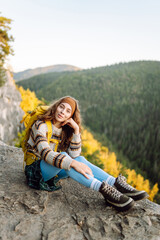 Young woman stands with her back to the edge of a rocky cliff, looking at the mountain landscape....