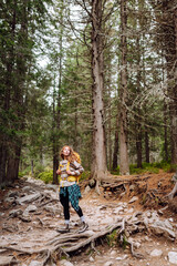 Obraz premium Young female hiker with hiking backpack walking up rocky path in lush green forest. Happy woman hiker enjoying nature on sunny day. Adventure, recreation concept. Active lifestyle.