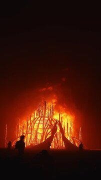A dome like wooden structure with spires burns intensely at night in a desert setting, with flames flickering and silhouettes of people watching.