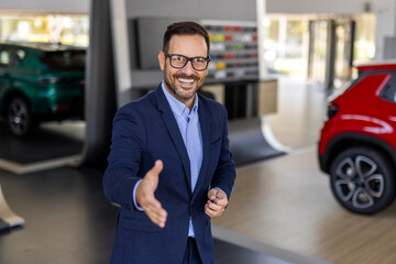Successful deal in a car showroom, young confident sales man reaching out for a handshake.