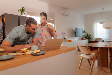 Fototapeta premium Young couple sitting together at a bright kitchen table, using a laptop while enjoying a healthy breakfast, savoring a peaceful morning in their modern home filled with warmth and connection