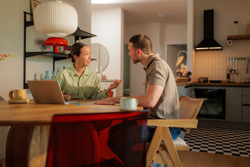 Young multi ethnic couple sitting at kitchen table discussing project details while using laptop and drinking coffee, enjoying working from home in modern apartment