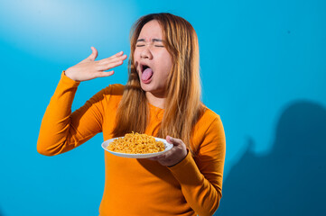 Asian woman in orange shirt with a disgusted expression, sticking out tongue while holding a plate of instant noodles, reacting negatively to the food against a blue background