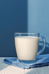 Clear glass mug containing fresh milk resting on napkin, positioned atop white wooden surface with blue background