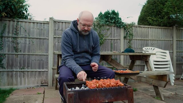 Man grilling marinated meat skewers over hot coals in backyard. Male preparing barbecue kebabs seated near rustic wooden picnic table. Guy cooking outdoor meal surrounded by simple patio and greenery