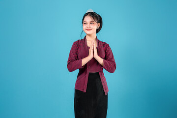 Smiling young Asian woman in traditional striped lurik attire with hands in greeting gesture. Her...