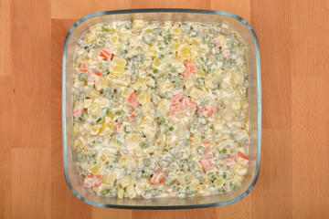 A glass dish filled with creamy Croatian-style French salad with visible diced vegetables and mayonnaise sits on a wooden surface, viewed from above