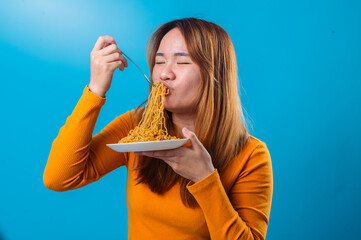 Asian woman in orange shirt eagerly slurping a forkful of instant noodles with eyes closed, showing...