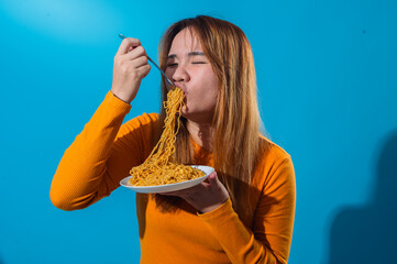 Asian woman in orange shirt eagerly slurping a forkful of instant noodles with eyes closed, showing enjoyment and hunger, in front of a clean blue background