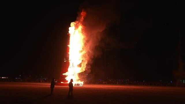 A dynamic scene of a wooden structure engulfed in flames at night, with flickering fire, billowing smoke, and silhouettes of observers in a desert setting.
