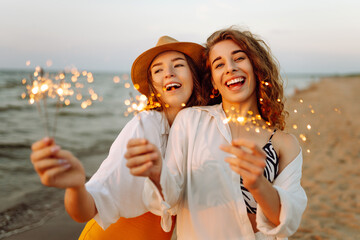 Beautiful girlfriends with sparklers joyfully enjoying the moment on the sunset beach. Two young friends spending time together, enjoying the lights outdoors. Concept of celebration, freedom.