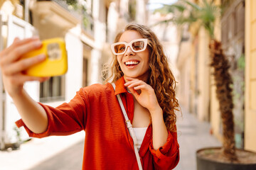 A smiling tourist in stylish clothes and with a phone walks along the sunny streets of the city. The woman takes a selfie and blogs with her phone outdoors. Blogging, travel concept.