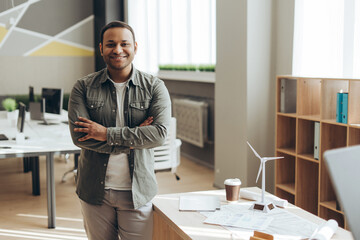 Indian engineer smiling with crossed arms in the office next to a wind turbine model