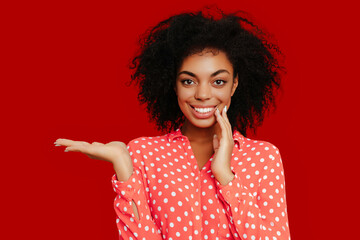 African American woman with natural curly hair, wearing a coral pink polka dot shirt, gesturing with one hand, presenting something and touching her face, posing against a bold red background.
