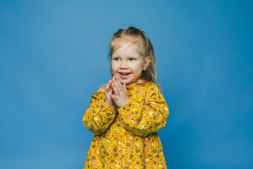 Little blonde girl in yellow dress smiling and clapping her hands. blue background.