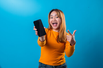 Asian woman in orange shirt with surprised expression pointing at a blank smartphone screen against a vibrant blue background in a promotional studio setup