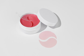 A white mockup jar with pink hydrogel eye patches and a spatula on a white isolated background....