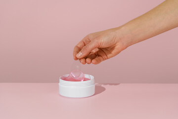 Female hand lifting with spatula pink under-eye hydrogel patch over white cosmetic jar, with pink...