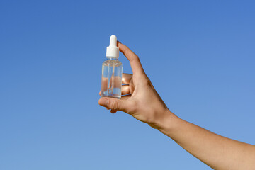 Female hand holding a transparent glass jar of serum with a pipette on a blue sky background. Concept of aesthetics and beauty care products, smoothing wrinkles and moisturizing the face.