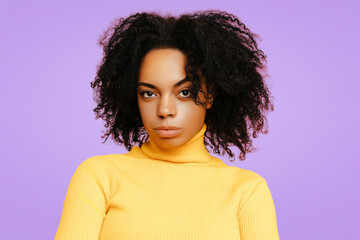 Confident African American woman with natural curly hair, wearing a bright yellow turtleneck sweater, posing with a serious, intense expression against a soft purple background.