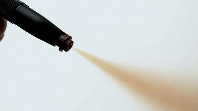 Close-up profile shot of the nozzle tip of an airbrush spray tan gun emitting a fine, even mist of bronzed tanning solution against a clean white backdrop