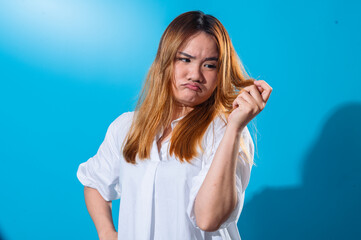 Fototapeta premium Asian woman in a white shirt looking at her hair with a concerned and displeased expression, examining split ends or dryness, standing against a solid blue background in studio light