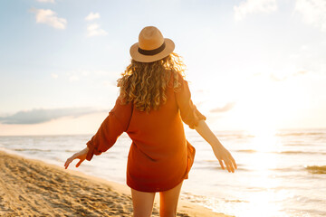Back view of a young woman with curly hair feels free on the seashore. Young woman walks on the...