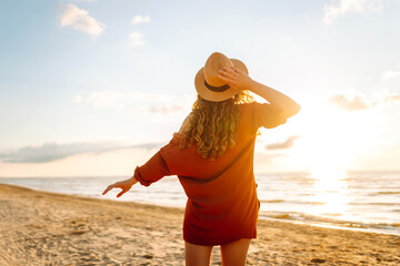 Back view of a young woman with curly hair feels free on the seashore. Young woman walks on the beach at sunset. Concept of relaxation, enjoyment, freedom.