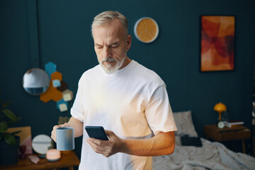 Elderly man with gray hair holding coffee cup while checking smartphone in cozy indoor setting...