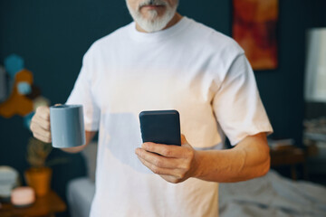 Elderly man holding smartphone and cup while standing in living room. Focused on device and enjoying moment in comfortable home environment