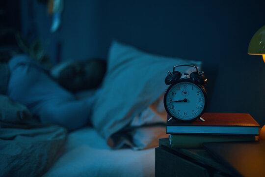 Person peacefully sleeping in bed with alarm clock and lamp on bedside table. Soft dim light creating calm and restful atmosphere