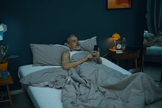 Middle-aged man with a beard lying in bed and using smartphone in a modern bedroom setting. Room decorated with plants and a bedside lamp providing a cozy atmosphere