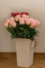 A beautiful bouquet of light pink and red roses is artfully arranged in a tall, rectangular pastel vase, showcased on a dark wooden floor in a bright indoor setting