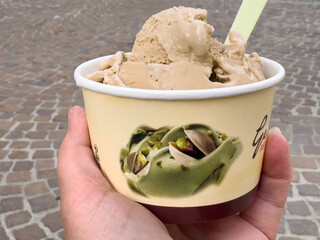 Hand holding coffee flavored gelato in cup with pistachio image on cobblestone street