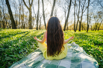 A woman with long dark hair does meditation in a sunny park in a green meadow with flowers, relaxation and yoga