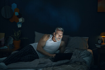 Senior man sitting in bed, illuminated by laptop screen, deeply engrossed in his work while...