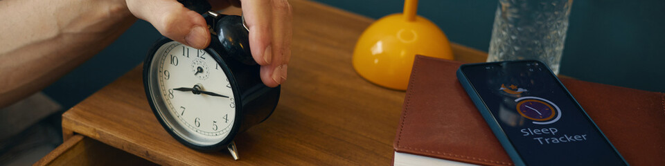Close-up of hand adjusting alarm clock on bedside table while another hand uses smartphone sleep...
