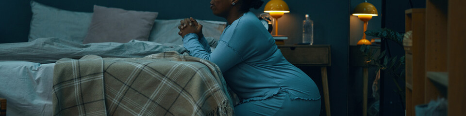 Woman kneeling beside bed, hands clasped in prayer, in a cozy, dimly lit bedroom setting with soft...