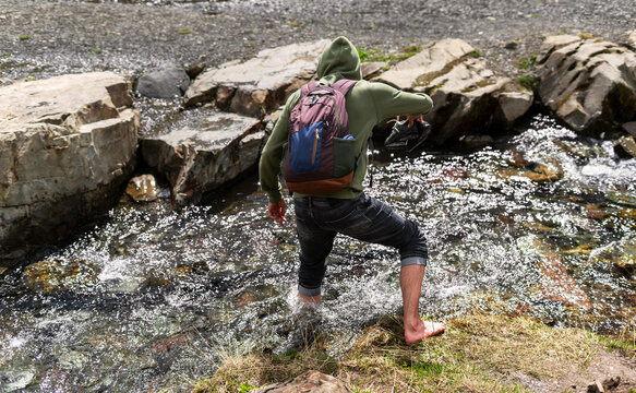 Young person wearing hoodie and backpack crosses rocky stream barefoot, holding shoes in hand Outdoor adventure and exploration in nature emphasizing self-reliance and balance, hiking and walking