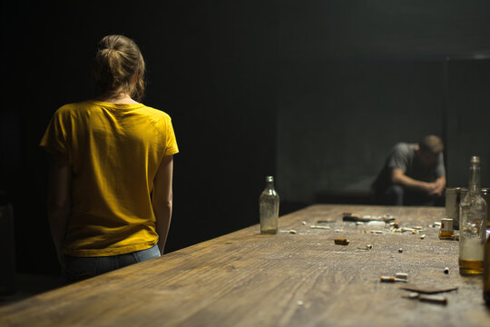 Woman in yellow shirt turns away from man slumped over table strewn with drug paraphernalia and empty bottles, depicting despair and addiction
