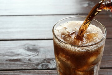 Pouring refreshing cola into glass at wooden table, closeup. Space for text