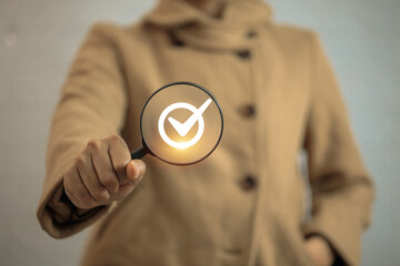 A person in a brown coat is holding a magnifying glass with a glowing checkmark icon inside,...