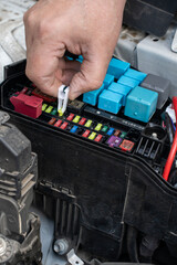 Hands are shown holding a blue fuse while standing over the engine compartment of a car.