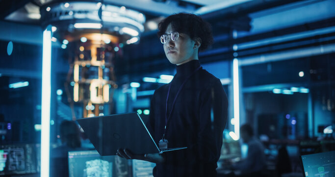 Asian Software Developer Using a Laptop to Monitor the Work of a Quantum Computer, Focusing on Data Flow, Compute and Hardware Stability. Futuristic Dark Setting with Advanced Technological Office
