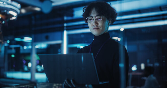 Asian Software Developer Using a Laptop to Monitor the Work of a Quantum Computer, Focusing on Data Flow, Compute and Hardware Stability. Futuristic Dark Setting with Advanced Technological Office - Powered by Adobe