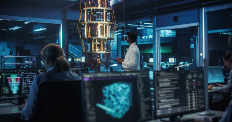 Young Indian Scientist Inspecting a Quantum Computer, Using a Tablet to Implement Complex Software Systems. Foreground with Artificial Intelligence Models on Office Screens