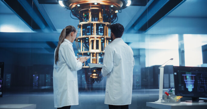 Young Caucasian Female Team Leader and Male Indian Scientist Talking in a Research Center with a Futuristic Quantum Computer Device. Specialists in White Coats Standing with Their Backs to Camera